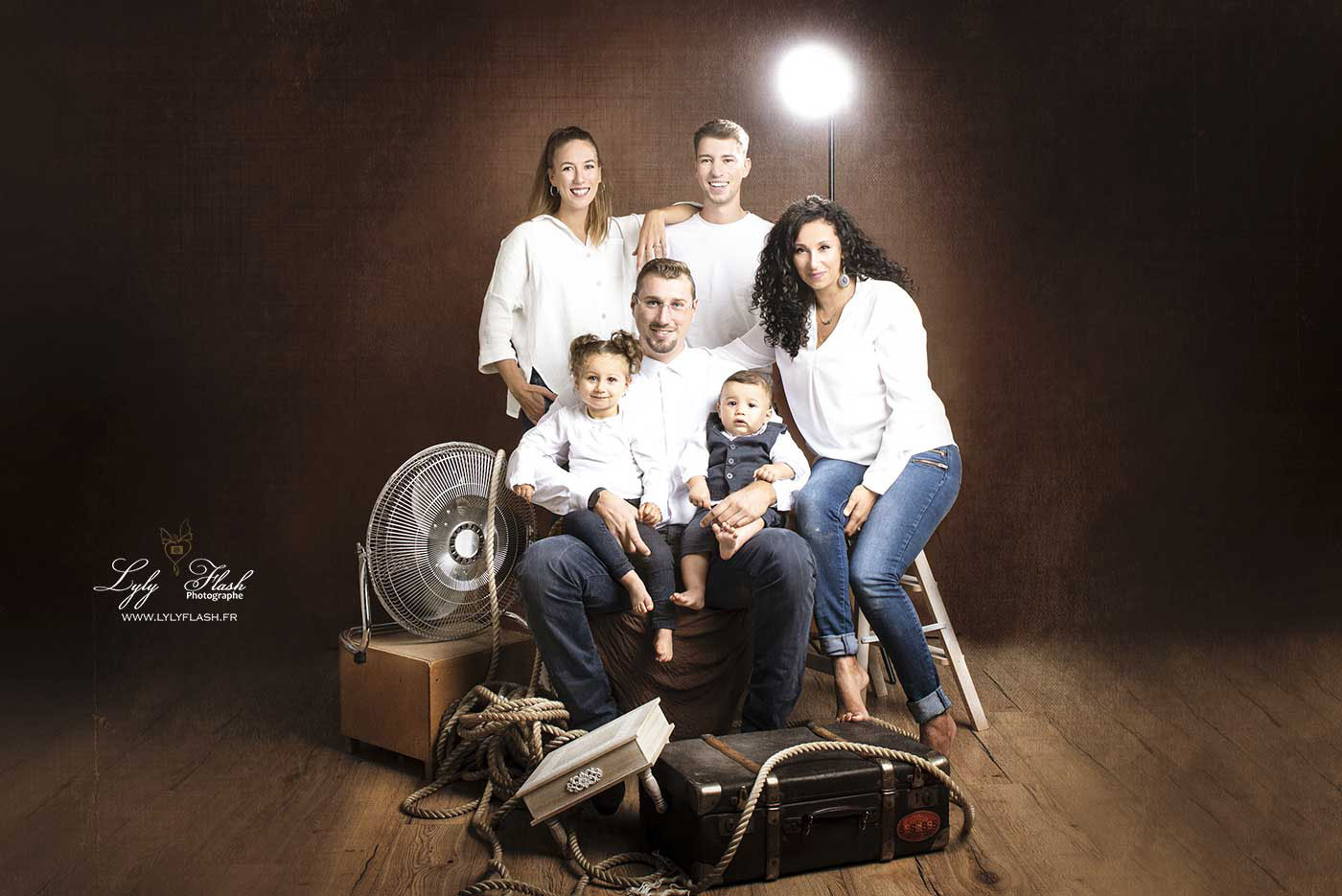 Dans cette magnifique photo de famille au style mi-vintage, mi-moderne, l’ambiance industrielle du studio apporte une touche à la fois brute et élégante. Les murs en béton texturé, les éclairages suspendus et les accessoires métalliques se mêlent à un mobilier vintage, comme un vieux canapé en cuir usé et une horloge rétro en arrière-plan. Cela crée un décor unique et raffiné. Le papa, habillé d'une chemise à carreaux et d’un pantalon en toile, est assis avec assurance sur le canapé, tenant leur bébé dans ses bras. Le bébé, tout souriant, porte une petite salopette vintage qui se marie parfaitement à l'esthétique rétro de la scène. La maman, vêtue d'une robe fluide avec des tons doux, est debout à côté, une main posée délicatement sur l’épaule de sa petite fille, qui porte une robe en dentelle blanche rappelant les anciens clichés de famille. À leurs côtés, le parrain et la marraine complètent le tableau, leurs tenues élégantes combinant des touches modernes et rétro : un costume avec des bretelles pour lui et une robe à motifs floraux vintage pour elle. Tous sourient chaleureusement, l’atmosphère est conviviale et naturelle, et l’union des générations est palpable dans ce cliché intemporel. Les lumières douces et les ombres bien placées apportent une sensation de profondeur et d’intimité, tout en mettant en valeur les expressions sincères et les liens forts qui unissent cette famille et leurs proches.