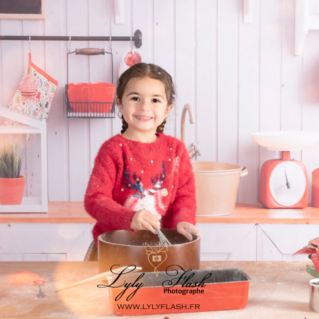 À La Garde, une petite fille avec un bonnet de Noël trop grand aide à étaler de la pâte à biscuit sur la table de la cuisine du Père Noël, le sourire aux lèvres.