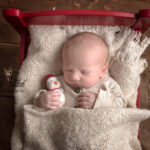 photographie d'un bébé qui dort dans un lit rouge Noël avec un bonhomme de neige comme doudou. La photographie célèbre sa naissance le 24 décembre avec cette belle mise en scène