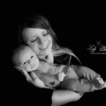 photographie noir et blanc, une jeune maman tient son nouveau-né dans ses bras près de son visage. Elle a un sourire qui explique tout son bonheur. photographie sur fond noir en studio réalisée près de Brenon par la photographe Lyly Flash