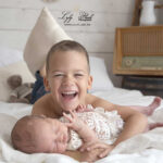 Un grand frère heureux en plein fou rire de bonheur a tenir sa petite sœur à la naissance, une photographie réalise dans le home studio comme a la maison du studio photo de la photographe Lyly flash pour cette famille de Vinon-sur-Verdon