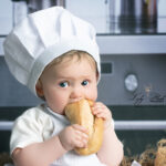 photo d un bébé de 1 an habillé en petit cuisinier dans une cuisine moderne qui mange une bonne baguette a la Française de Paris